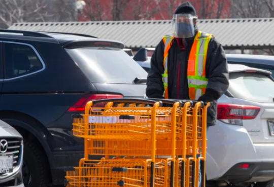 Worker wearing face shield pushing grocery carts in store parking lot