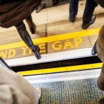 View from above of people walking over "Mind the Gap" writing on floor of London Underground while exiting the tube