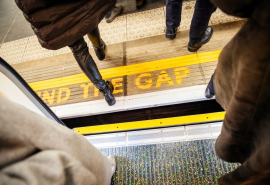 View from above of people walking over "Mind the Gap" writing on floor of London Underground while exiting the tube