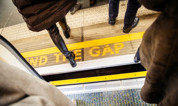 View from above of people walking over "Mind the Gap" writing on floor of London Underground while exiting the tube