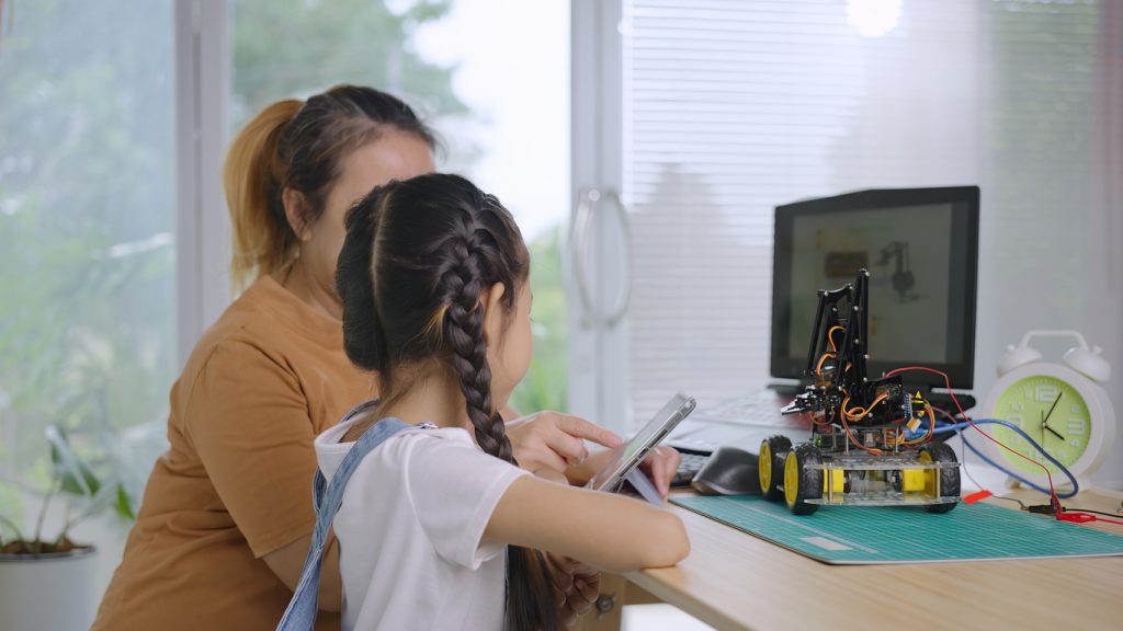 Mom and young daughter pictured from behind, building robot together on desk.