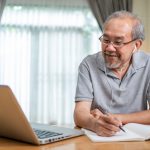 Older man working on laptop at home and writing in notebook