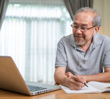 Older man working on laptop at home and writing in notebook