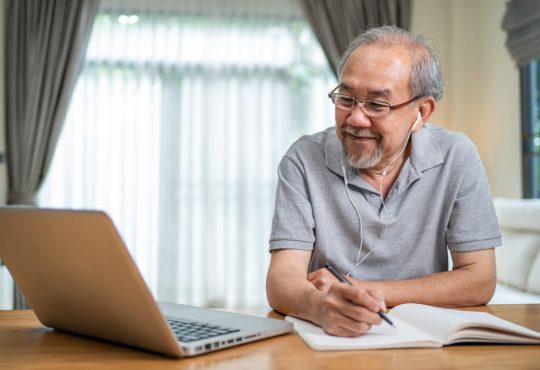 Older man working on laptop at home and writing in notebook