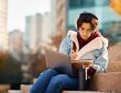 Young woman writing in notebook with laptop on lap sitting on stairs outside on university campus