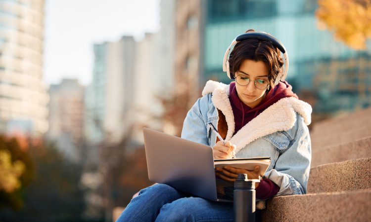 Young woman writing in notebook with laptop on lap sitting on stairs outside on university campus