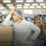 Woman wearing white lab coat and hairnet in factory leaning against stack of boxes with head in hand