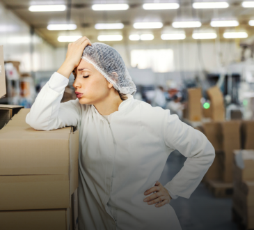 Woman wearing white lab coat and hairnet in factory leaning against stack of boxes with head in hand