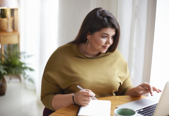 Woman working on computer and writing in notebook at home