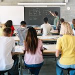 Students seen from back of classroom while teacher teaches at blackboard