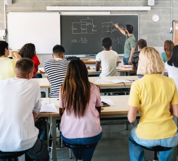 Students seen from back of classroom while teacher teaches at blackboard