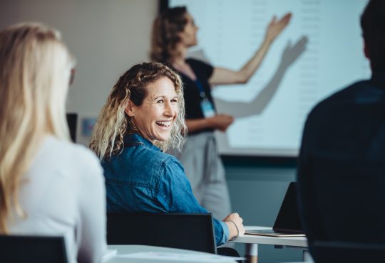 Rear view of a woman sitting in audience looking back at a colleague and smiling during a conference.