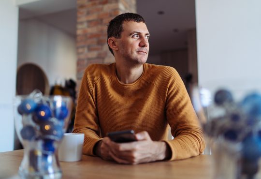 A man wearing yellow sweater sitting in the kitchen and looking through window while using smartphone