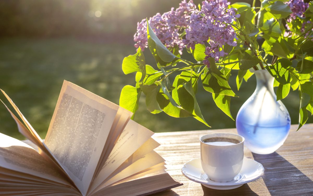 Open book sitting on table outdoors beside teacup and vase with purple lilacs