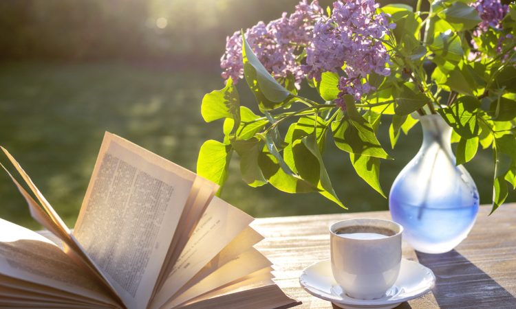 Open book sitting on table outdoors beside teacup and vase with purple lilacs