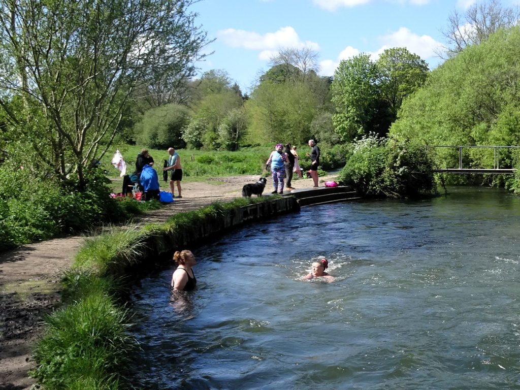 People swim in a river while others stand and walk on a pathway nearby