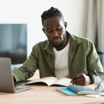 Black man working on laptop and taking notes in notebook