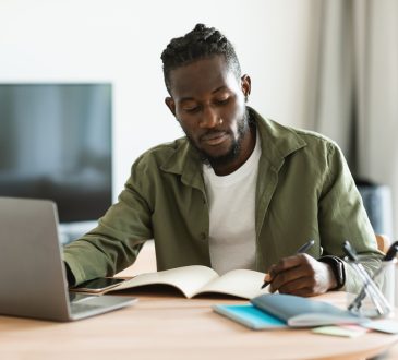Black man working on laptop and taking notes in notebook