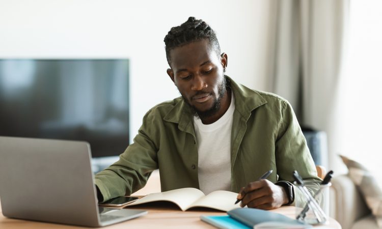 Black man working on laptop and taking notes in notebook