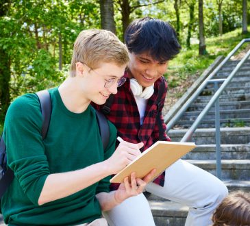 Two teens smiling while looking at notebook, leaning on railing against backdrop of trees on campus