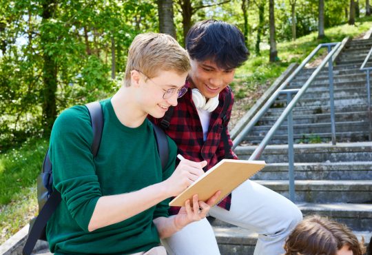 Two teens smiling while looking at notebook, leaning on railing against backdrop of trees on campus
