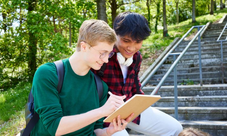 Two teens smiling while looking at notebook, leaning on railing against backdrop of trees on campus