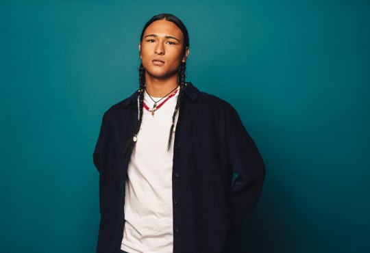 Indigenous man standing in front of a blue wall facing the camera.