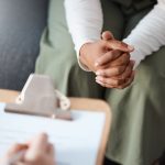 Closeup of person sitting with hands clasped in lap across from person writing on clipboard