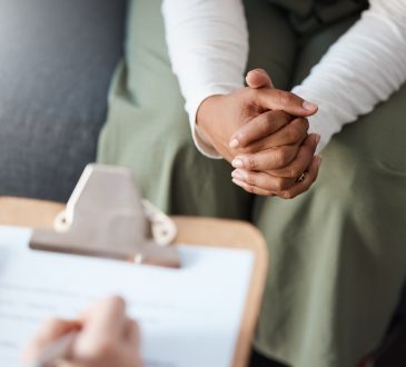 Closeup of person sitting with hands clasped in lap across from person writing on clipboard