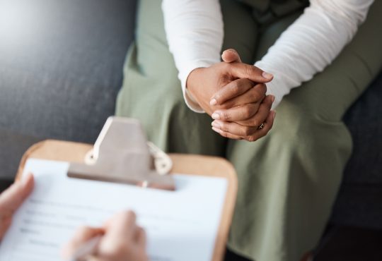 Closeup of person sitting with hands clasped in lap across from person writing on clipboard