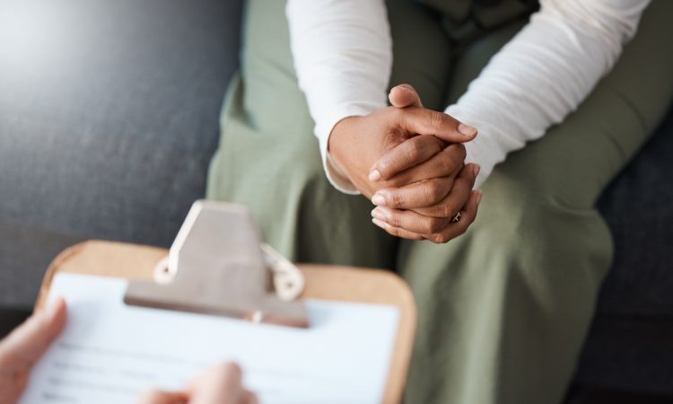 Closeup of person sitting with hands clasped in lap across from person writing on clipboard