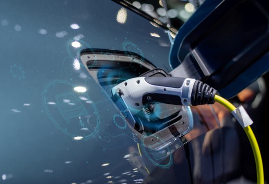 The image shows an electric vehicle charging at a charging station, with a close-up of the charging plug inserted into the vehicle and digital graphics overlaying the scene, suggesting technology and connectivity.