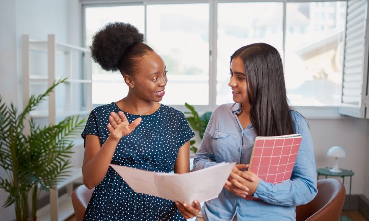 Two women standing next to each other and facing one another. they are talking to each other with one woman holding a piece of paper in front of them.