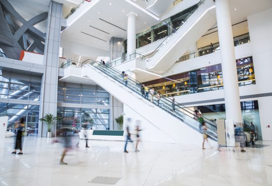 The image shows the interior of a modern, spacious shopping mall with multiple levels connected by escalators, where people are moving about, creating a sense of activity and movement.