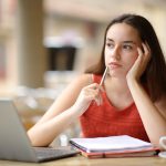 Student sitting at laptop thinking with hand holding side of head and pen raised to mouth