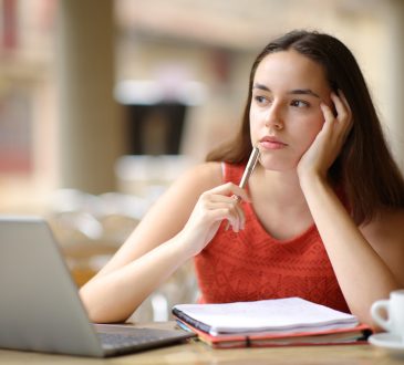 Student sitting at laptop thinking with hand holding side of head and pen raised to mouth