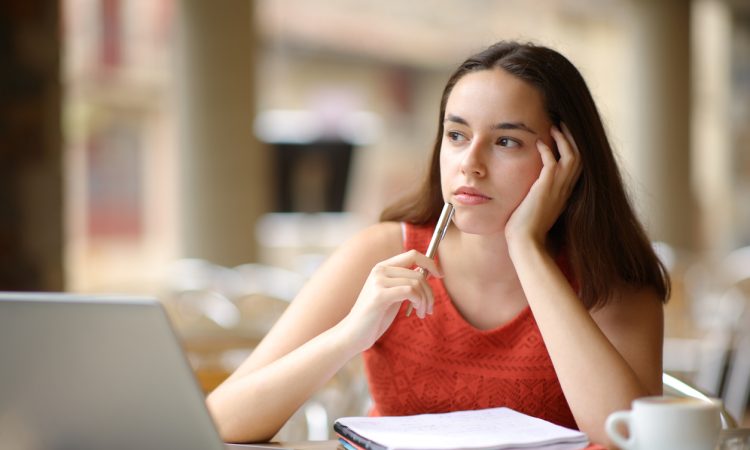 Student sitting at laptop thinking with hand holding side of head and pen raised to mouth