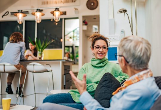 A young woman with glasses and a green turtleneck, smiling and engaging in conversation with an older woman in a modern, brightly lit living space.