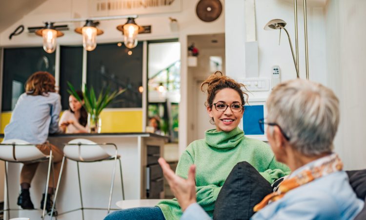 A young woman with glasses and a green turtleneck, smiling and engaging in conversation with an older woman in a modern, brightly lit living space.