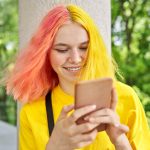 Teen with yellow and pink hair smiling and looking at phone outside