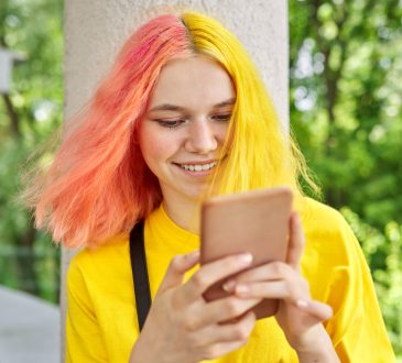 Teen with yellow and pink hair smiling and looking at phone outside