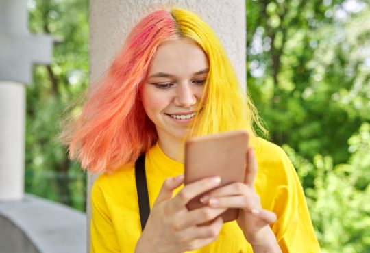 Teen with yellow and pink hair smiling and looking at phone outside