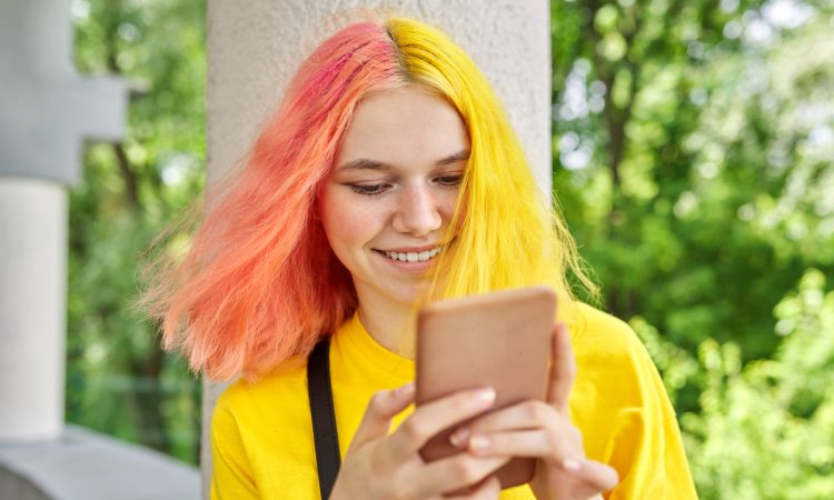 Teen with yellow and pink hair smiling and looking at phone outside