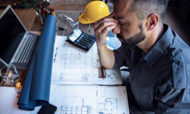 Photo of a stressed out construction worker with his eyes closed and hand to his face sitting over a desk with blue prints.