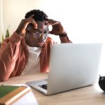 The image shows a young man sitting at a desk, focused on his laptop with a frustrated or concentrated expression, hands touching his head.