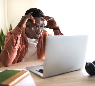 The image shows a young man sitting at a desk, focused on his laptop with a frustrated or concentrated expression, hands touching his head.