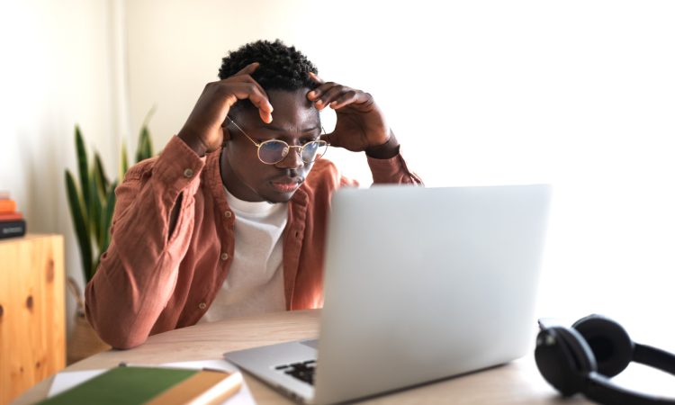 The image shows a young man sitting at a desk, focused on his laptop with a frustrated or concentrated expression, hands touching his head.