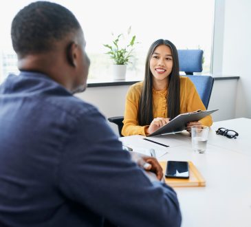 The image shows a professional setting where a smiling interviewer, holding a clipboard, is engaged in a conversation with a candidate across the table.