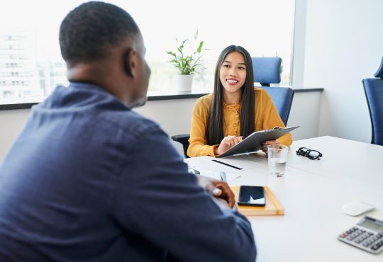 The image shows a professional setting where a smiling interviewer, holding a clipboard, is engaged in a conversation with a candidate across the table.