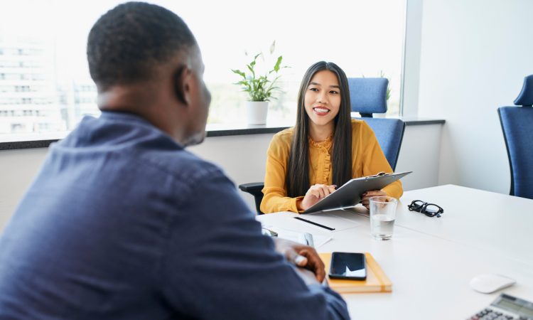 The image shows a professional setting where a smiling interviewer, holding a clipboard, is engaged in a conversation with a candidate across the table.